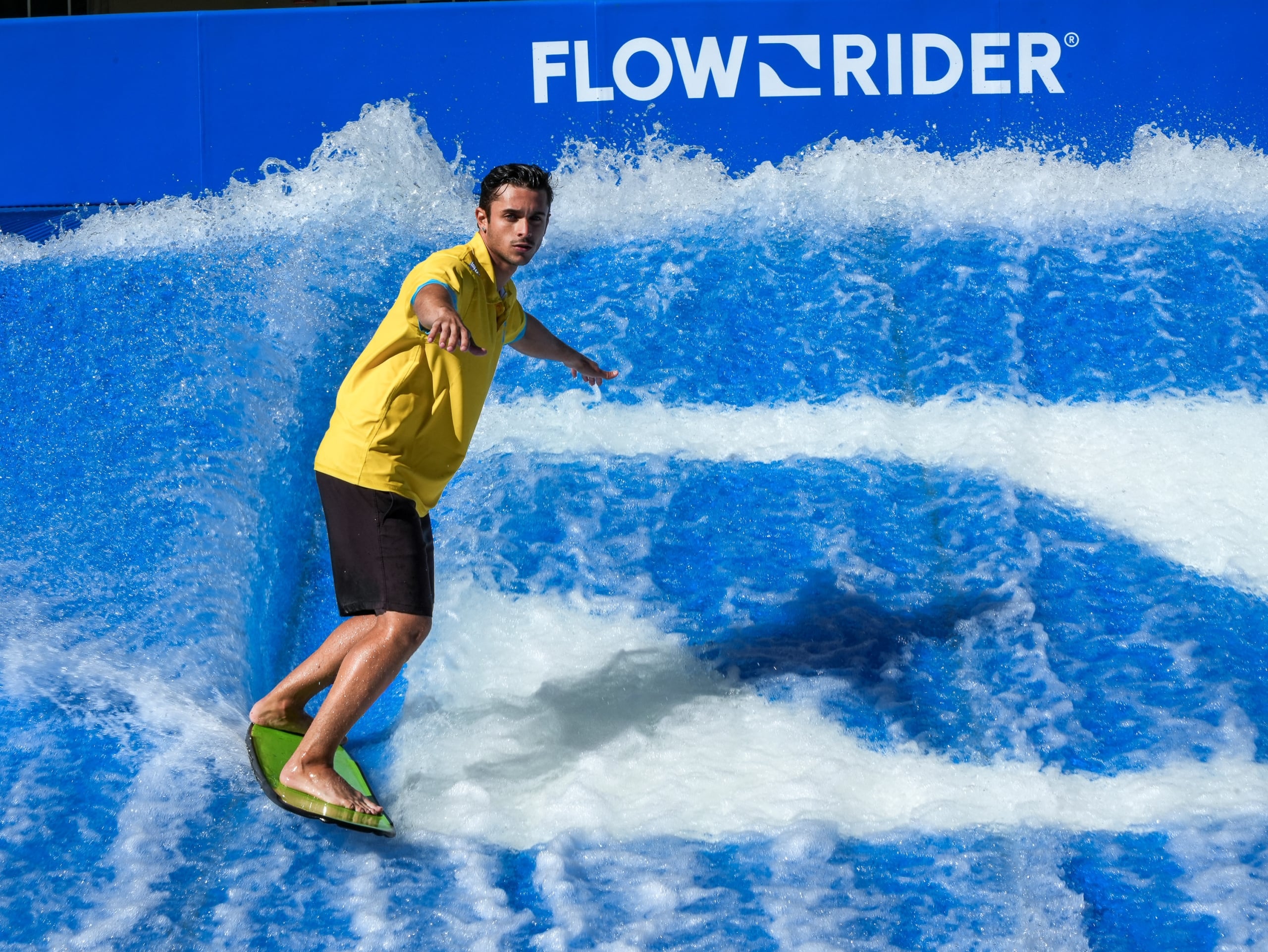 A male dressed in a yellow polo and black board shorts rides the FlowRider wave generator