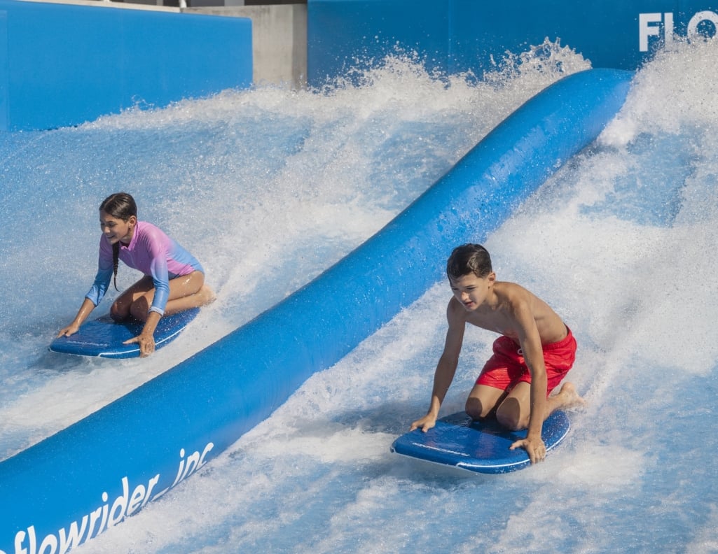A boy and girl ride the Flow Rider knelt atop their surf boards