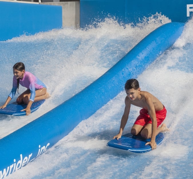 A boy and girl ride the Flow Rider knelt atop their surf boards