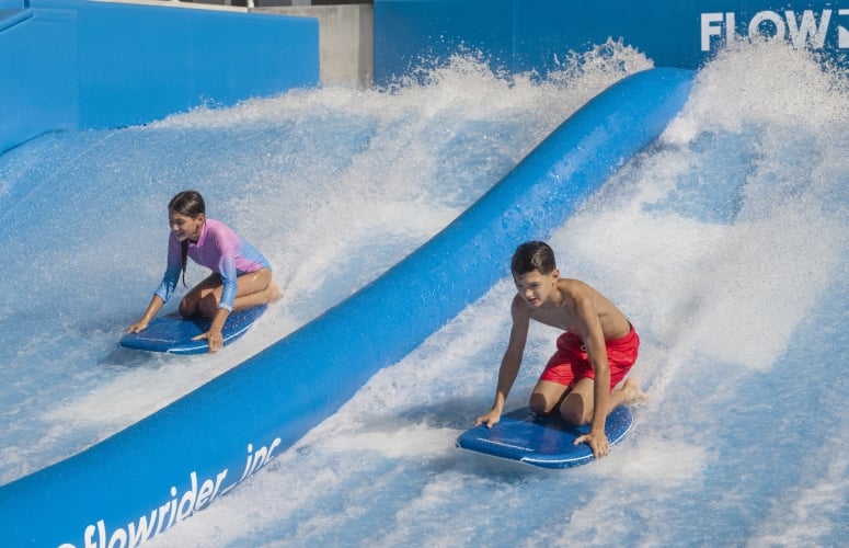 A boy and girl ride the Flow Rider knelt atop their surf boards
