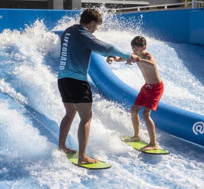 A lifeguard and boy hold hands for balance as they surf the Flow Rider attraction