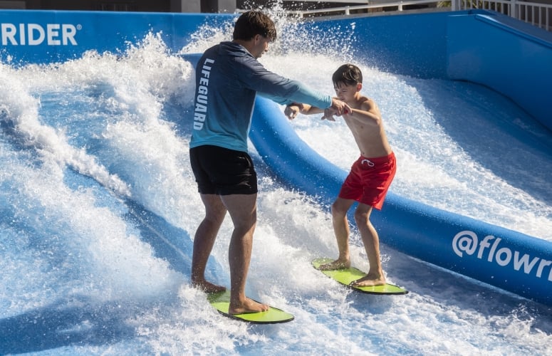 A lifeguard and boy hold hands for balance as they surf the Flow Rider attraction