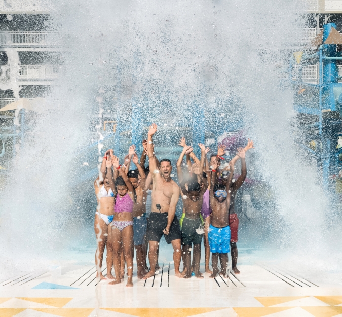 A large group of individuals stand under The Tipping Bucket as water pours over them, at Surf and splash Waterpark