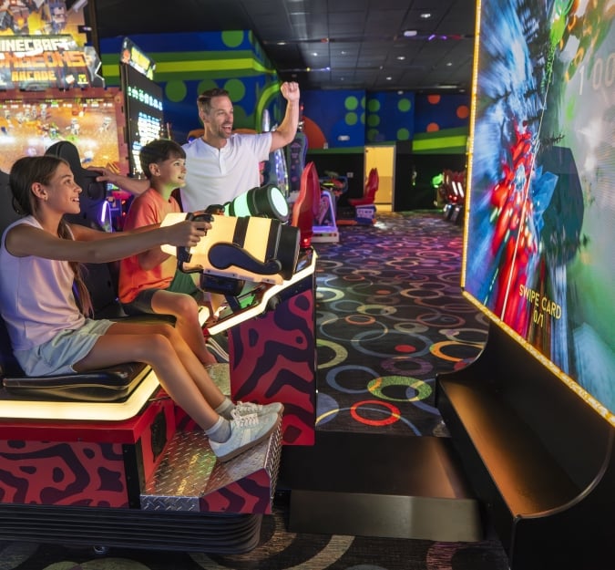 Children playing an arcade game together as dad cheers from the sidelines.